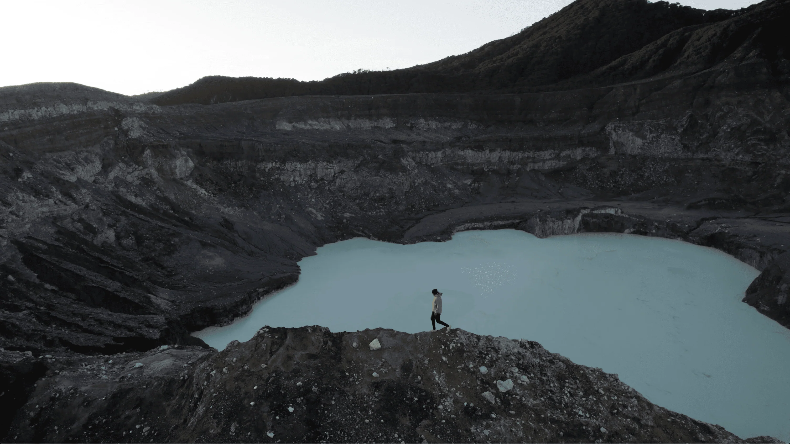 A person in a yellow jacket walking along the edge of the Poas volcano crater, standing near the milky lake, with rocky slopes and forested hills in the background.