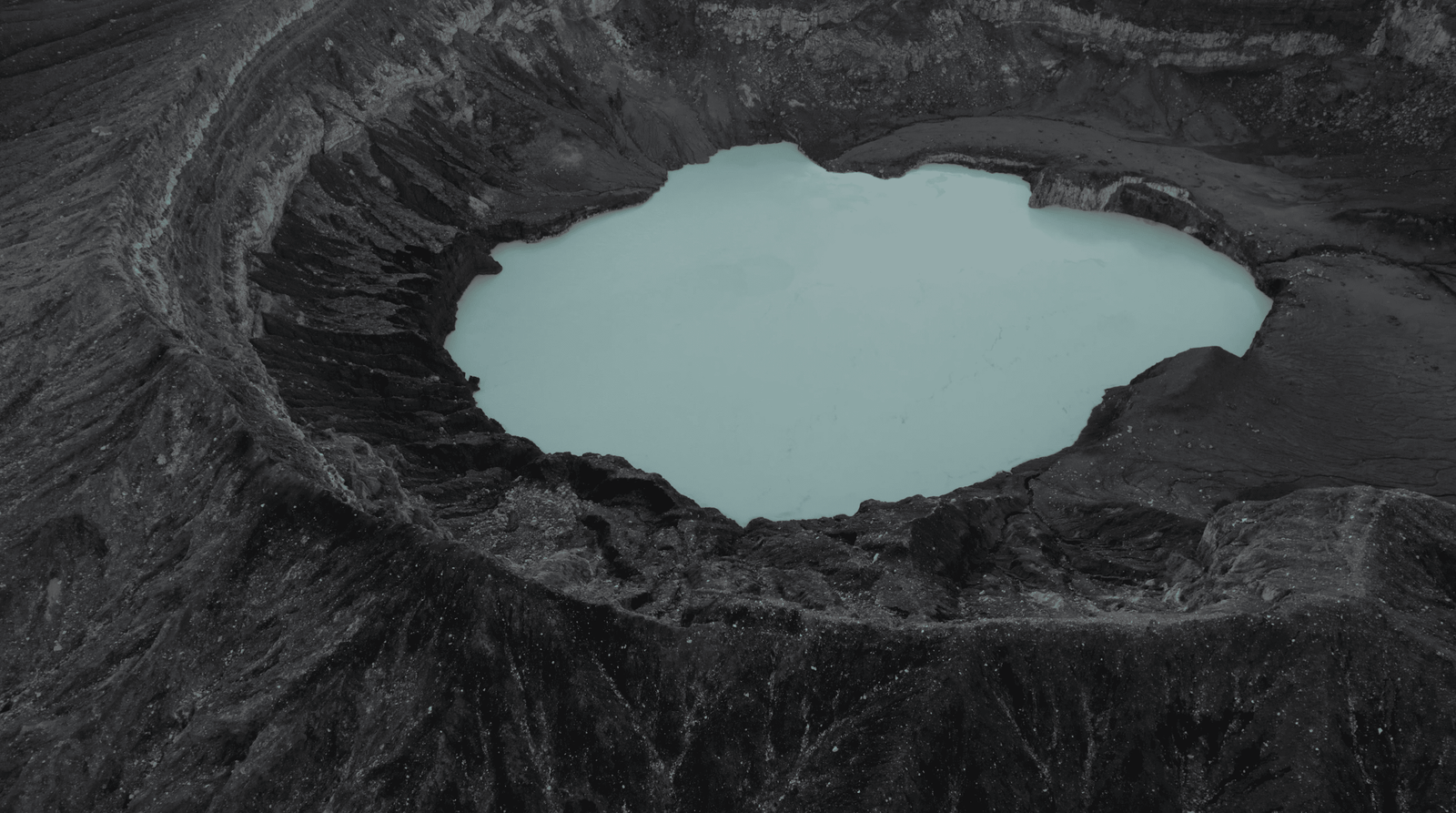Poas Volcano: Worth a Visit in 2026? 13 Aerial view of Poas volcano crater with a milky turquoise lake surrounded by dark, rocky terrain and lush green hills in the background.