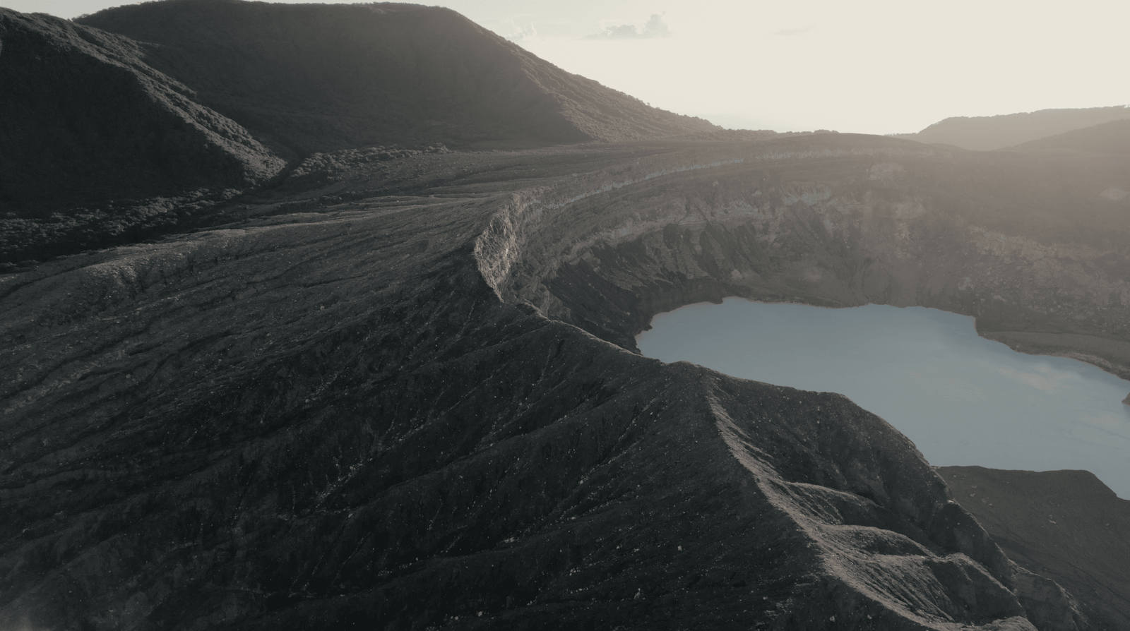 Poas Volcano: Worth a Visit in 2026? 7 Aerial view of Poas volcano crater with a milky turquoise lake surrounded by dark, rocky terrain and lush green hills in the background.