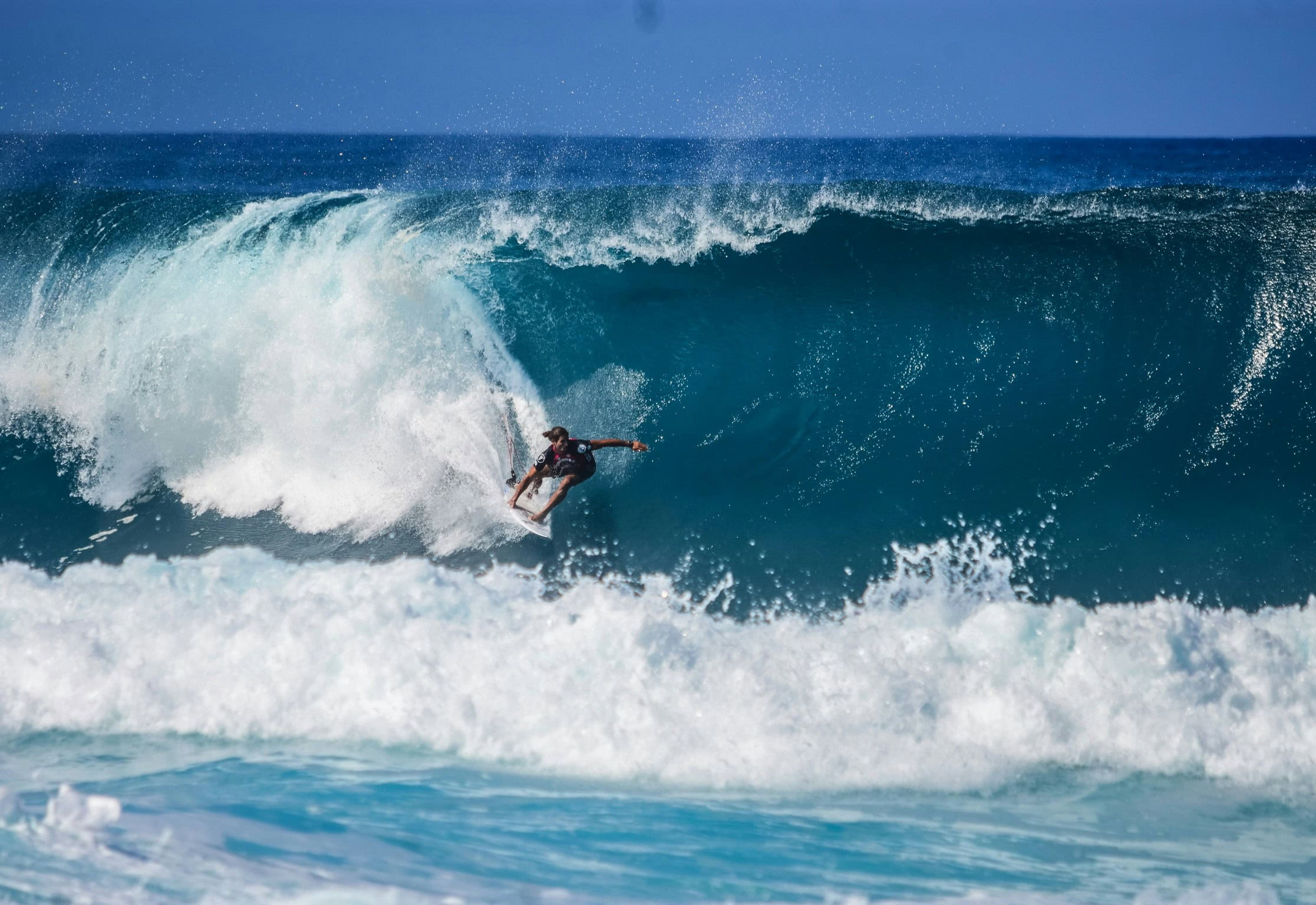 A surfer rides deep inside the barrel of a massive turquoise wave, skillfully leaning into the curve with spray trailing behind the board. The powerful ocean swell crashes around him under a clear blue sky.