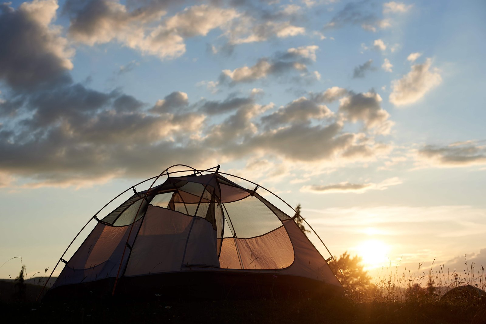 Camping Tips For Beginners in 2026! 4 Camping at daybreak. The tent stands under a blue sky with clouds in the rays of evening sunlight on a blurred background of trees and in the distance of the mountains