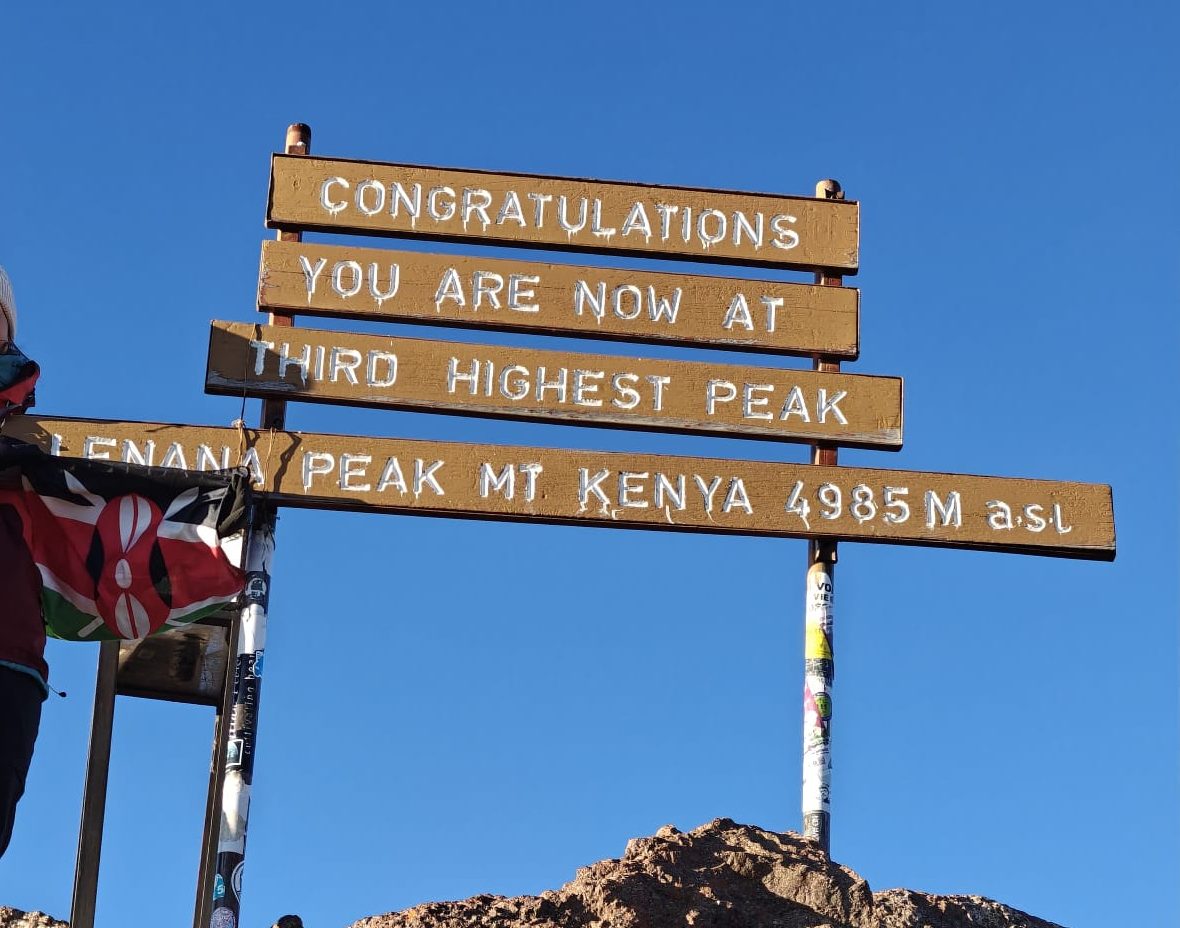 Panoramic view of a hiker at the summit of Mount Kenya at Point Lenana, standing next to a wooden sign marking the third highest peak of Mount Kenya, with clear blue sky overhead.