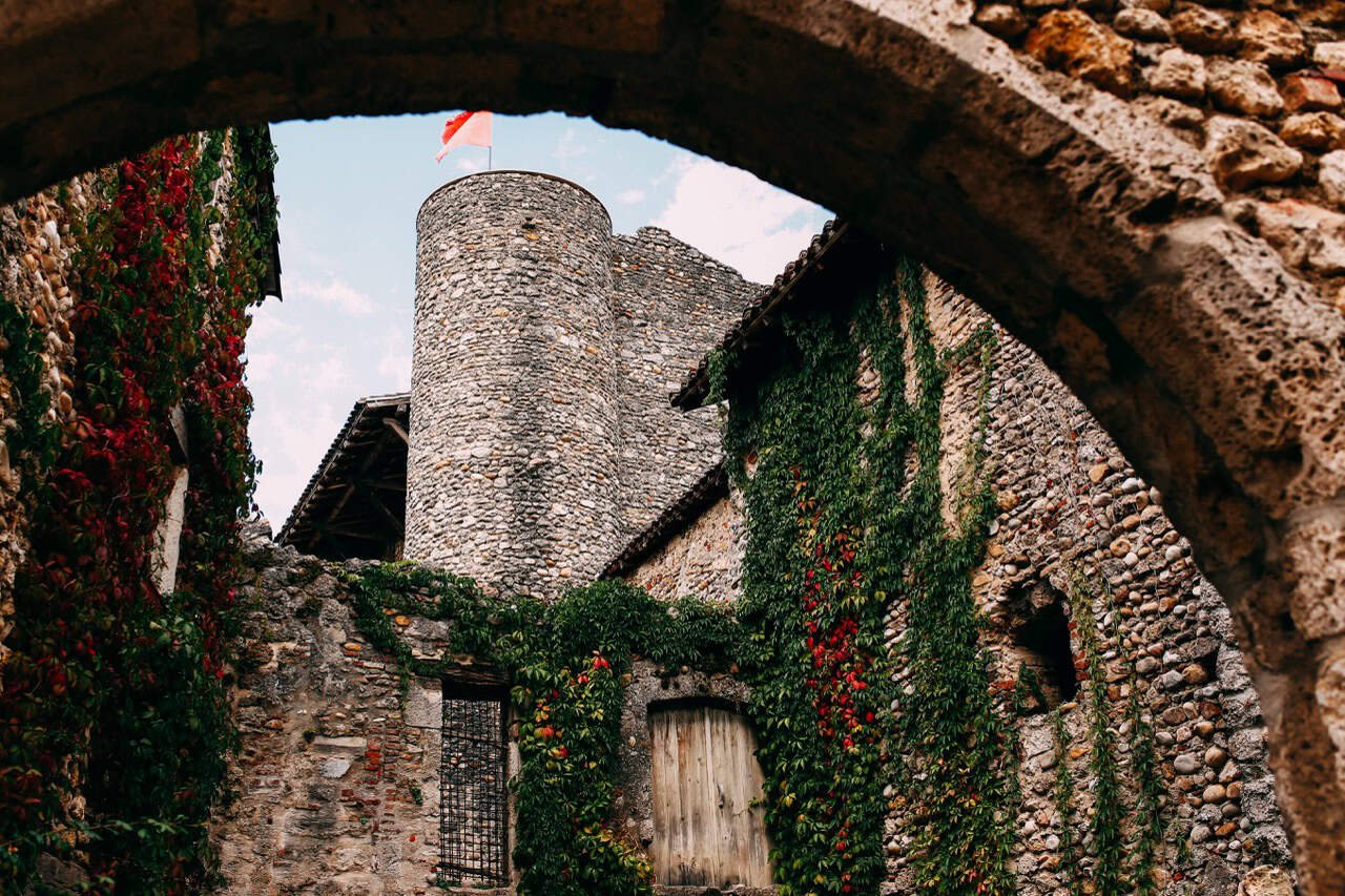 7 Charming Medieval Villages in France (2026 Expert Guide) 7 Ancient stone castle çin Perouges with lush green vines and red flowers, viewed through an archway, showcasing historic architecture and scenic beauty. Perfect for exploring medieval castles and European landmarks.
